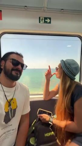 A man and a woman are riding on a train with a view of the ocean. The man is sitting on the left side of the frame, and the woman is sitting on the right side of the frame. The man is wearing a white t-shirt with a graphic on it, sunglasses, and a necklace. He has dark hair and a beard. The woman is wearing a denim hat, sunglasses, and a dark tank top. She has long, red hair. They are both looking out the window at the ocean. The man gives a thumbs-up to the camera. The woman holds her hand up. The train is moving and the ocean is visible through the window. There is an emergency exit sign visible at the top of the frame. The woman is holding a phone in her hand.