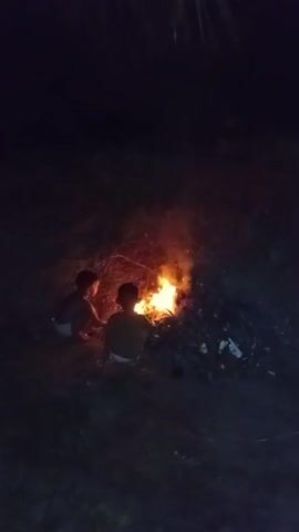 The video shows two young children sitting around a small bonfire at night in Buketteukuh, Indonesia. The fire illuminates their faces and bodies, casting a warm glow on the surrounding darkness. The children appear to be interacting with the fire, possibly playing or warming themselves. The setting seems to be an outdoor area, with dry leaves and twigs forming the base of the bonfire. The time of day is clearly nighttime, indicated by the darkness and the illuminated fire. No text is visible in the video. The overall mood is one of childhood innocence and simple enjoyment of a natural phenomenon.