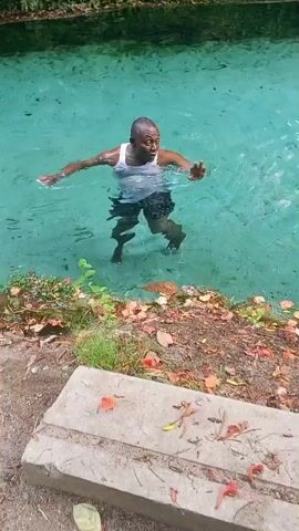 A short, handheld video shows a dark-skinned man standing in a clear, light blue pool of water. He is wearing a white tank top and dark shorts. The man has a shaved head. He appears to be talking while looking up. The pool is surrounded by lush green vegetation and fallen red petals. A concrete block sits near the edge of the pool.