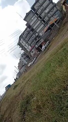 The video shows an exterior, medium shot of a grassy slope beside a street in a town. Several buildings are visible in the background, including a tall apartment building and smaller shops. Power lines crisscross the sky, which is partially covered by clouds. Several cars are parked along the street, and two people are walking on the grassy area. A blue truck is parked further down the street. The camera is angled, giving a tilted perspective of the scene.