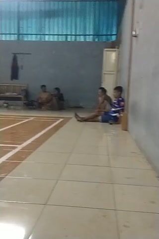 The video shows a group of people playing badminton indoors on a wooden court. The court is marked with white lines. The lighting is somewhat dim, with bright lights visible in the background. Several individuals are actively engaged in playing the sport, with racquets in hand and in motion. One person in a blue shirt is in the foreground, seemingly in the middle of a game. Other players in dark shirts are also visible on the court. The overall environment suggests a badminton facility.