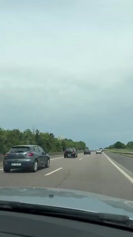 The video shows the perspective of someone driving on a highway with multiple lanes. There are several cars in front, and trees line the side of the road. In the distance, a large, white industrial structure can be seen against a cloudy sky. The road is asphalt with clear lane markings.