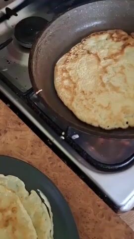 The video shows a close-up of a person making crepes. The camera pans over a skillet on a gas stove, where a crepe is being cooked. The crepe is golden brown with some darker spots. The person then flips the crepe with a spatula, revealing the cooked side. The crepe is then placed on a plate with other crepes. The video ends with a close-up of the stack of crepes.