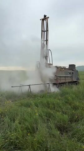 A drilling rig is operating in a grassy field under an overcast sky. The rig is emitting a large amount of white smoke or steam. Two individuals are visible near the base of the rig, one standing and observing, the other appears to be operating or assisting with the machinery. Metal pipes are positioned in front of the rig, supported by stands. The field is covered in tall green grass and some yellow flowers. The overall scene suggests an industrial or exploratory operation taking place outdoors.