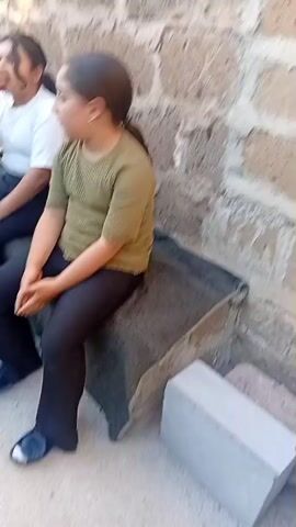 TWO YOUNG GIRLS ARE SITTING ON A STONE BENCH OUTSIDE. THE GIRL ON THE LEFT IS WEARING A WHITE SHIRT AND BLACK PANTS AND HAS TWO LIGHT BLUE BALLOONS IN HER LAP. SHE IS LOOKING TO THE LEFT AND APPEARS TO BE EATING SOMETHING. THE GIRL ON THE RIGHT IS WEARING A GREEN SHIRT AND BLACK PANTS. SHE IS LOOKING FORWARD WITH HER HANDS CLASPED IN HER LAP. THE WALL BEHIND THEM IS MADE OF ROUGH STONE. A GREY CONCRETE BLOCK IS VISIBLE IN THE FOREGROUND TO THE RIGHT.