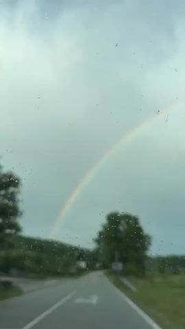 The video depicts a rainy day scene from the perspective of someone inside a vehicle, looking through the windshield. The windshield has raindrops on it. The road ahead is visible, marked with a white arrow indicating the direction of travel. A rainbow arcs across the sky in the distance. Green trees and other vegetation line the side of the road. There is a road sign on the right. Music can be heard in the background.