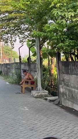 A shirtless man sits on a plastic chair on the side of a paved walkway. He wears dark shorts and a baseball cap. A pair of sandals rests on the ground near his feet. He is looking down and appears to be tying his shoes or adjusting something on his feet. The walkway is bordered by a low concrete wall on the left and a taller concrete wall on the right. Behind the left wall, there is a fence and some vegetation. Behind the right wall, there are trees and dense foliage. A light pole stands behind the man and the chair. The sun is shining, casting shadows.