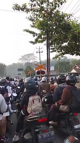 The video shows a long line of motorbikes stuck in traffic at a railway crossing. The riders are mostly wearing helmets, and many have backpacks. A train is visible passing through the crossing, and the red lights signal that it is unsafe to cross. The environment appears to be a road with trees and utility poles. The sky is overcast, suggesting it might be morning or late afternoon. The mood seems to be one of patience as the riders wait for the train to pass.