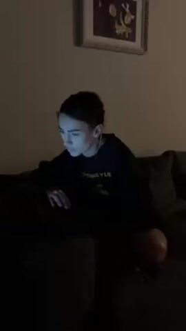 A young white woman with dark hair pulled back and wearing a black t-shirt with a yellow graphic, is sitting on a dark couch in dim lighting, using a laptop. She has wired earbuds in her ears. A framed picture hangs on the wall behind her. The light from the laptop illuminates her face as she looks at the screen.