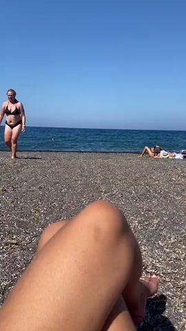 The video pans across a black pebble beach under a clear blue sky. In the foreground, a person's legs are visible, resting on a beanbag chair. In the mid-ground, a woman in a black bikini walks towards the camera. Further back, several people are sunbathing and relaxing on beanbag chairs and under a wooden canopy. The ocean is visible in the background, with gentle waves lapping the shore. The overall scene depicts a relaxed beach atmosphere on a sunny day.
