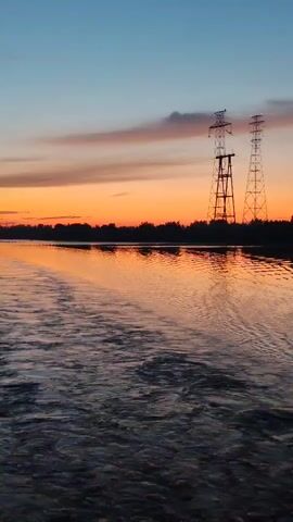The video shows a wide river at dusk. The sky is a gradient of blue and orange, with thin, wispy clouds. Two tall, metal power line towers stand on the far bank of the river, silhouetted against the sunset. The water is rippled, suggesting movement, and reflects the colors of the sky. The scene is peaceful and serene.