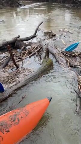 The video shows two men in life vests, dressed in outdoor clothing, standing near a muddy riverbank littered with fallen branches. One man is holding a shovel and smiles at the camera. The camera pans to show a murky river with many fallen tree branches partially submerged in the water. The men are then seen using the shovel and their hands to move the branches in the river. One man is wearing an orange waterproof jacket and the other is wearing a dark blue one. Both men are also wearing orange life vests over their jackets. The scene is set on a cloudy day near a river surrounded by bare trees.