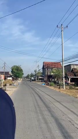 The video shows a perspective from a vehicle moving down a rural asphalt road. The sky is clear and blue with some wispy clouds. Telephone poles with numerous wires run along the side of the road. On the left, there are houses with red tile roofs, trees, and piles of straw or hay. On the right, there are more houses, some with weathered stone walls and red tile roofs. In the distance, a few motorcycles are visible on the road. The road has tire marks on it.