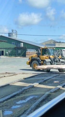 The media file captures an outdoor scene at what appears to be an industrial or agricultural facility in Verijärve, Estonia. The primary subject is a large yellow construction vehicle, likely a wheel loader, parked on a paved area. The vehicle is facing to the right of the frame, with its bucket raised. Behind the loader, other vehicles and buildings are visible, suggesting a working environment. One building is predominantly green with a corrugated metal exterior and several windows. Another structure, to the left and further back, is lighter in color and appears to be a warehouse or processing plant, possibly with solar panels on its roof.

The scene is set during the daytime, under a bright blue sky with scattered white clouds, indicating fair weather. The ground in the foreground is a mix of gravel and a rough concrete-like surface. The lighting suggests it is mid-day, with clear shadows cast by the objects.

There are no people clearly visible in the media file. The overall impression is one of industrial activity or preparation for work. The location cues, such as the architecture of the buildings and the type of vehicles, are consistent with a rural or semi-industrial setting in Estonia. No text is discernible in the media file.