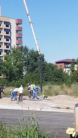 In a construction site scene, a concrete pump boom extends diagonally upwards from the right of the frame towards the left. Five construction workers in blue overalls and other work attire are gathered around the base of the boom. They appear to be operating shovels, rakes, and possibly a trowel, suggesting they are working with concrete or other construction materials. One worker in a yellow vest is visible among them. Behind the construction site, a multi-story apartment building with balconies and windows stands tall. Lush green trees and vegetation are situated between the construction area and the building. In the foreground, the edge of a paved road or sidewalk is visible, with some green grass and weeds growing nearby. A woman with short grey hair and a colorful polka-dot top, wearing blue jeans and carrying a red bag on wheels, stands looking at the construction activity from the right side of the road. The sky is clear and blue. The scene is captured in daylight, indicating an outdoor work environment.