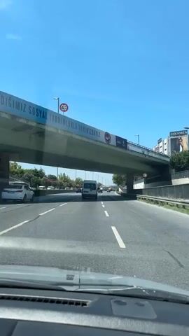 The video begins from the perspective of a driver inside a car, looking forward through the windshield. The car is on a highway, driving under an overpass. The overpass has a large banner on its side with Turkish text and a logo. A sign with "4.70" indicating height restriction is visible on the overpass. The highway has multiple lanes, and other vehicles, including cars and vans, are driving in the same direction. The weather is sunny with a clear blue sky. Streetlights are visible along the side of the highway, and trees and some buildings are seen in the background. The camera captures the road ahead, the vehicles in front and to the sides, and the overpass above. The video continues to show the car driving on the highway, passing under the overpass, and continuing its journey.
