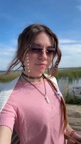 A woman with long brown hair, sunglasses, a pink shirt, and jewelry takes a selfie outdoors. She is standing on a paved path next to a body of water and some greenery. In the background, there are trees, buildings, and a boat. The sky is blue with some clouds.