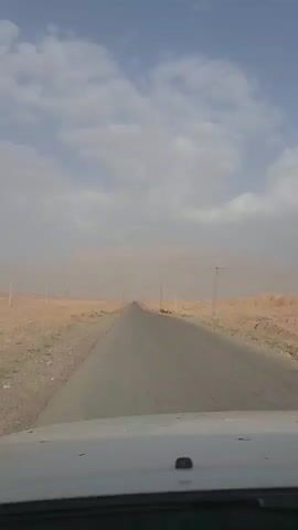 A car is driving on a straight paved road through a desert landscape. The road is surrounded by dry, sandy terrain with sparse vegetation. The sky is partly cloudy, with some patches of blue and scattered white clouds. In the distance, mountains are visible, partially obscured by dust or haze. Several utility poles with wires are spaced along the sides of the road. The view is from the perspective of someone inside the car, looking forward through the windshield. The interior of the car, specifically the dashboard and hood, is visible in the foreground. The overall impression is one of a journey through a remote, arid environment.
