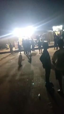 The video is a low-resolution, shaky, nighttime recording of an outdoor scene with multiple people present. The lighting is dim, with bright artificial lights illuminating parts of the background, possibly from stalls or streetlights. The ground appears to be paved or dirt. Silhouettes of individuals are visible, suggesting they are standing and some are in motion. The focus is poor, making it difficult to discern specific details about the people's clothing or faces. There are also some blurry shapes in the background that could be vehicles or structures. The overall impression is of a public gathering or street scene at night.
