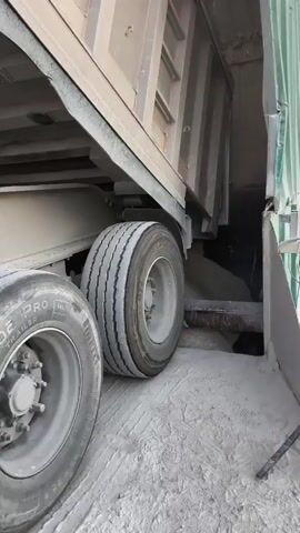 A close-up, low-angle shot shows the rear dual tires of a dump truck filled with a gray granular substance, possibly cement or aggregate. The truck bed is raised, revealing the material piled inside. The camera pans slightly to the right, focusing on the opening of the truck bed where the material is being discharged, showing a chute or conveyor system. The surrounding ground is also covered in the same gray substance, indicating a dusty, industrial environment, possibly a construction site or material processing facility. No people are visible.