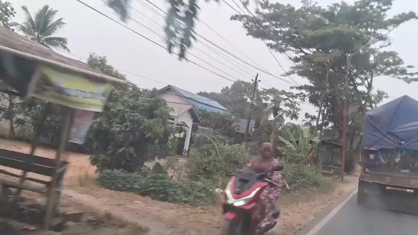 The video shows a view from a moving vehicle, looking out a window. The scene is rural, with houses and trees visible. There are power lines overhead. The road appears to be unpaved. The weather is overcast. The vehicle is moving from left to right in the frame.