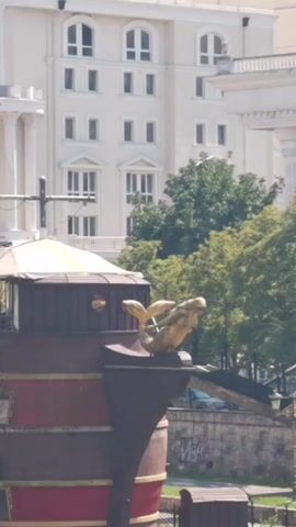 A close-up view of a decorative ship's prow with a golden mermaid figurehead is seen in the foreground. Behind it, a wide shot reveals a multi-story building with arched windows and a tree-lined embankment. The embankment is littered with trash, including plastic bottles and other debris. A person rides a bicycle along a paved path on the embankment. The water in the foreground is rippling.