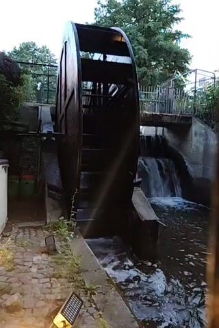 A water wheel with wooden paddles is partially visible on the left. Water flows over a concrete structure behind the wheel, creating a small waterfall. A metal fence with vertical bars is in the foreground, obscuring parts of the scene. A yellow sign with black lettering is attached to the fence. The environment appears to be outdoors with trees and greenery in the background. The lighting suggests it may be dusk or overcast.