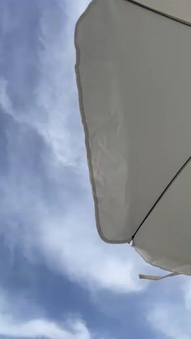 The video is an extreme close-up shot looking upwards at a white beach umbrella against a blue sky with scattered clouds. The umbrella is angled to the right, filling a significant portion of the frame. The edge of the umbrella is wavy and somewhat creased. The clouds are thin and wispy, streaking across the sky. The bottom left corner of the frame briefly shows a blurred pinkish-orange object. The video is static with no discernible movement other than slight shifts in light and shadow on the umbrella.