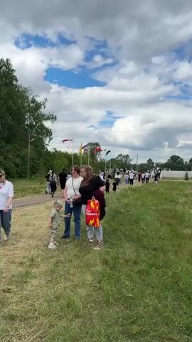 The video shows a large outdoor gathering of people in a grassy field under a cloudy sky. In the foreground, two women are standing on the left, and a woman with two children walks towards the right. More people are visible in the middle ground, walking and milling about. Further in the background, there are tents, flags, and trees. A crane is also visible in the distance on the left. The overall scene suggests a festival, fair, or community event.