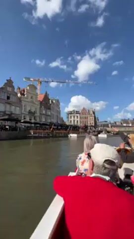 A group of people are on a boat traveling along a canal. They are looking at the buildings on either side of the canal. The buildings are old and have a variety of architectural styles. There is a construction crane in the background. The sky is blue and there are some white clouds.