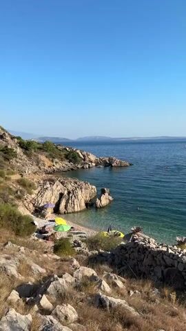The video pans across a rocky coastline on a sunny day. The ocean is a clear turquoise, with several boats and swimmers visible in the distance. In the foreground, there is a small pebble beach with a few colorful umbrellas and straw-covered sun shelters. The rocky terrain is covered with dry grass and some low-lying vegetation. The sky is a clear, bright blue.