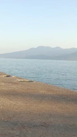 This is a landscape video that shows a body of water and a landmass in the distance, likely a mountain range under a clear sky. In the foreground, there is a dirt and gravel surface, and a pier extends into the water. Several people are gathered on the pier, some standing, some sitting, and some on motorcycles. A few dogs are also present on the pier and the land. The water appears calm and the lighting suggests it is either early morning or late afternoon.