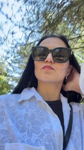 The video shows a woman with long dark hair and sunglasses, wearing a white button-up shirt over a black top. She is walking on a wooden boardwalk in a forest. The sun is shining through the trees. In the background, other people can be seen walking on the boardwalk. The woman is smiling and looking at the camera.