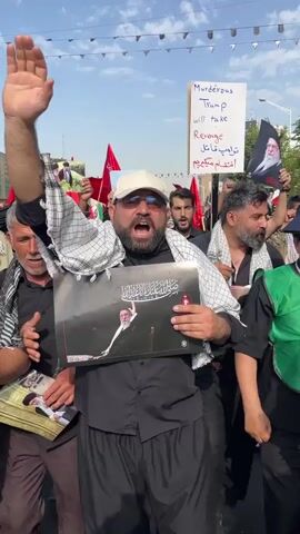 A group of men are gathered outdoors, with several holding flags and signs. One man in the foreground, wearing a black shirt, a white baseball cap, and a checkered scarf around his neck, is holding a large poster with an image of a man. He is raising his right hand in the air and appears to be shouting. Another man to his left, also wearing a black shirt and a checkered scarf, is raising his left hand and shouting. To the right of the man with the poster, another man in a black shirt and checkered scarf is holding a sign that reads "Murderous Trump will take Revenge" in English and Arabic. In the background, more people are visible, some holding red flags. The environment appears to be a street, with buildings and trees in the distance, and banners strung across the street overhead. The overall atmosphere seems to be one of protest or a rally.