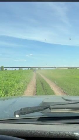 The video shows a view from inside a vehicle, looking out through the windshield. A dirt road curves through a green grassy field under a bright blue sky with some clouds. In the distance, a long, elevated structure, possibly a bridge or highway overpass, stretches across the horizon. The vehicle appears to be moving forward, as indicated by the slight blur and the changing perspective of the road. The dashboard of the vehicle is visible in the foreground.