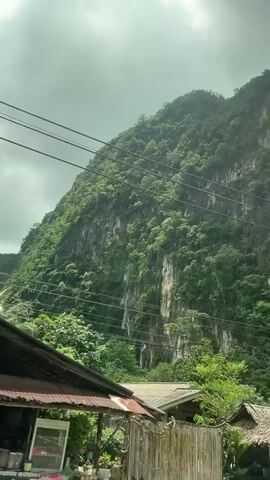The video shows a landscape with a large, green, vegetated mountain. There are several power lines running across the scene. In the foreground, there are some simple structures that appear to be houses or small businesses, constructed with wood and corrugated metal. There is also lush green vegetation, including trees and tall grass. The sky is overcast with clouds. The camera is moving, suggesting it is from a vehicle.