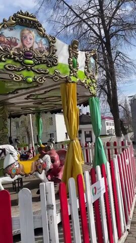 This video shows a stationary carousel in an outdoor setting, likely a park. The carousel is brightly colored with green, gold, and white accents, featuring animated characters on its upper panels, including what appears to be Rapunzel and characters from "Masha and the Bear." Several horse figures, primarily white and brown, and one elephant figure, are visible on the carousel's platform. Yellow and green drapes are tied around some of the carousel's support poles, suggesting it might be closed or awaiting operation.

A red and white picket fence encloses the carousel. In the background, there are bare trees, white architectural pillars, and several buildings, indicating an urban or suburban environment. The sky is partly cloudy, and the lighting suggests it's daytime. No people are visible in the video, giving the scene a quiet and deserted feel. A small sign on the fence has a heart icon, but the text beneath it is illegible. The location is Urussu, Russia.