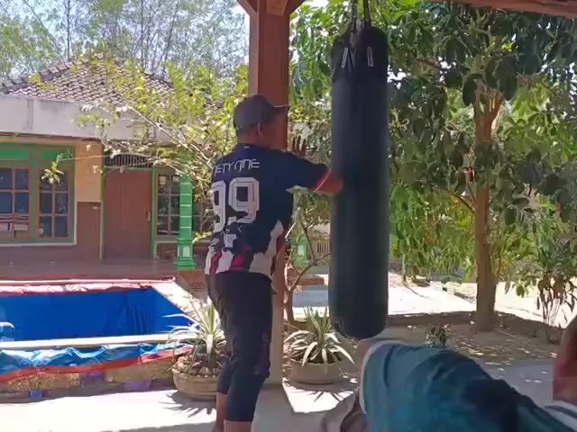 A man wearing a baseball cap and a t-shirt with the number 99 on the back is standing next to a black punching bag. He appears to be in the process of punching the bag. The man is standing outdoors under a wooden awning, with a house and trees in the background. There is a blue tarp spread out on the ground to the left of the man, and a few potted plants are visible. Another person is partially visible in the lower right corner of the frame, sitting and looking towards the punching bag. The weather appears to be sunny.