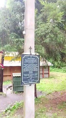 This is a video of a nature setting, likely a park or forest, located in Staryi Pokrov, Russia. The camera pans across the scene, capturing various elements of the natural environment and some man-made structures.

In the foreground, a rough, dirt path winds through lush green grass and under the shade of tall, mature trees. The foliage is dense, indicating a natural, possibly wild setting. Sunlight filters through the canopy, suggesting it is daytime, likely morning or afternoon, and the weather appears to be overcast or the lighting is diffused.

A prominent feature is a tall, concrete pole situated in the middle of the frame. Attached to this pole is a dark, rectangular plaque with a decorative silver border. At the top of the plaque, a small, dark cross is affixed. The plaque itself contains text, written in a language that appears to be Cyrillic. The text is difficult to read clearly due to the video's resolution, but it seems to be informational, possibly historical or memorial in nature.

Behind the pole, a low, wooden structure with a yellow roof is visible. To the left of this structure, a bird feeder is attached to a tree, with a cluster of bright red flowers adorning the tree trunk below it. Further to the left, a darker green, louvered structure stands, resembling a small shed or enclosure.

The overall impression of the scene is one of tranquility and natural beauty, with a touch of historical or religious significance suggested by the plaque and the cross. The setting appears to be a quiet, perhaps secluded area within a forested landscape. There are no people visible in the video, and the activity is limited to the passive observation of the environment.