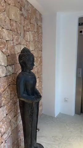 The video pans across a hallway. On the left, a dark statue of Buddha stands against a stone wall. To the right of the statue, a white wall leads to a stainless-steel elevator door. Below the elevator door, a small electrical outlet is visible. Further to the right, there are several bottles of water and some watermelons. The camera then moves to an alcove in a white wall, where two wooden statues are displayed on a shelf, lit by spotlights. The video ends with a view of the sea and trees through a window, with a security camera mounted on the wall above the window.