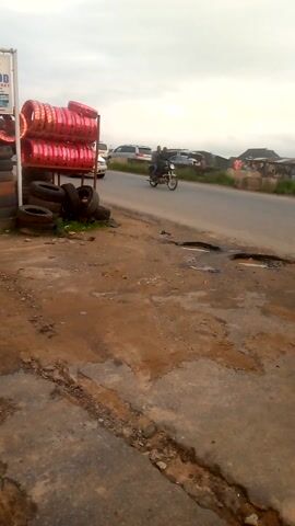 The video shows a street scene in Ore, Nigeria, on an overcast day. In the foreground on the left, a tire shop displays new red tires wrapped in plastic on a metal rack, alongside stacks of used black tires on the ground. A partially visible white sign with blue text reads "N GOD" and "MARKET," likely advertising the shop.

The right side of the foreground features a cracked asphalt and dirt shoulder with potholes and small puddles. A paved road runs through the middle ground, where a white SUV is parked, and motorcycles drive by. In the background, other vehicles and buildings are visible.

From 0:05 to 0:06, three pedestrians walk from right to left on the dirt shoulder: a man in a white and red striped shirt, followed by a woman in a red shirt, and another woman in a blue patterned dress. The overall scene depicts a busy roadside environment with commercial activity and pedestrian traffic.
