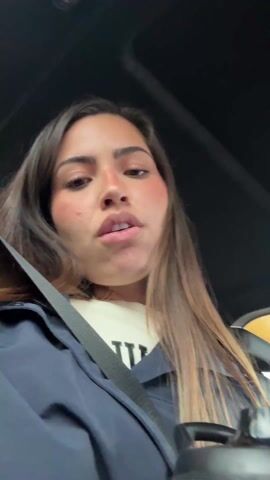 The video shows a woman in the driver's seat of a car, wearing a seatbelt, a dark blue jacket, and a white sweater. She has long brown hair and appears to be talking directly to the camera. The interior of the car is visible, with a black dashboard and a water bottle in the center console. Music is playing in the background. The woman's expressions change as she speaks.