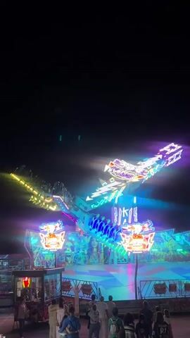 This vertical video captures a thrilling amusement park ride at night in Torres Vedras, Portugal. The ride, a large, multi-armed contraption adorned with vibrant, flashing lights in neon blues, purples, and yellows, dominates the frame. At the center of the ride, a long arm extends upwards and outwards, with seats for riders attached to the ends. These seats are also lit, creating a dazzling spectacle against the dark night sky.

Below the main spinning mechanism, there are smaller, spinning platforms, also brightly illuminated. The base of the ride features colorful geometric patterns and a large image of a woman's face, adding to the lively atmosphere. A crowd of people is gathered at the bottom of the frame, watching the ride in action. They appear to be enjoying the evening, with many looking up at the ride. Some individuals are closer to the ride's entrance, while others are further back. The lighting suggests it is nighttime, and the clear sky indicates fair weather. The overall mood is one of excitement and entertainment.