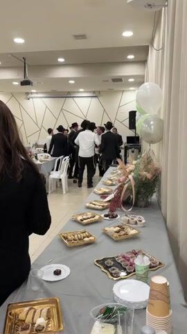 This media file captures a lively celebration, likely a wedding or Bar Mitzvah, taking place indoors at a venue in Kiryat Malachi, Israel. A group of men, predominantly dressed in black suits and hats, are dancing and celebrating in the background. A woman is visible in the foreground, her back to the camera, wearing a black sweater. A young girl in a light blue dress and red hair accessory stands near the dancing men.

The scene is set up with a long table laden with an assortment of pastries, cakes, and desserts, arranged in golden trays and platters. A floral arrangement with pink and white flowers adds a decorative touch. Three balloons, two white and one green, are tied to a stand near the table, suggesting a festive occasion. Disposable cups and plates are also visible.

The lighting indicates it is likely daytime or an evening event with bright artificial lighting. The ambiance appears joyful and energetic, with the dancing men clearly engaged in celebration. The architectural style of the room, with its geometric patterned wall and modern ceiling fixtures, suggests a contemporary event space. No visible text is present in the media file.