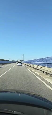 A vertical, first-person perspective video shows a car driving on a clear, sunny day. The vehicle is on a multi-lane highway that appears to be a bridge, indicated by the presence of large suspension towers and a blue barrier fence along the side. Several other cars are visible on the road ahead, including a white car in the immediate foreground. The sky is a vibrant, cloudless blue. The view is from the driver's seat, with the dashboard and windshield wipers visible at the bottom of the frame.