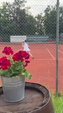 A woman in a white tennis outfit serves a tennis ball on a clay court, viewed through a chain-link fence. In the foreground, a potted plant with red flowers sits on a wooden barrel. The background shows trees and a banner on the fence behind the court.