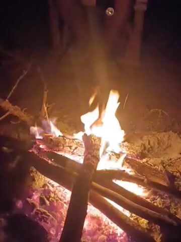 The video shows a close-up shot of a small campfire burning at night. The flames are bright and flickering, illuminating the surrounding sticks and sand. In the background, there are blurry figures of people sitting near the fire. Someone speaks in the background in Romanian, but what is being said is unintelligible. The setting seems to be outdoors.