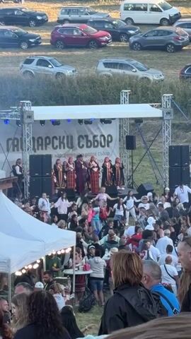 A high-angle, wide shot shows an outdoor festival with a stage set up in the background. A group of performers dressed in traditional attire stand on the stage. Below the stage, a large crowd of people are gathered, many of whom are seated on the grass. In the upper portion of the frame, a row of parked cars is visible on a grassy hillside. The lighting suggests it is daytime, likely late afternoon or early evening. The overall atmosphere appears to be festive and community-oriented.