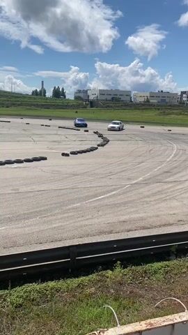 Two cars, one white and one blue, are drifting on an asphalt track. The white car is in the foreground, drifting to the right, while the blue car is behind and to the left, also drifting. Tires are laid out as markers on the track. In the background, there are buildings and a radio tower. The sky is blue with scattered white clouds. The track appears to be an outdoor circuit.