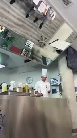A chef wearing a white uniform and hat stands behind a stainless steel counter in a commercial kitchen. He is preparing food and talking, gesturing with his hands. Behind him, there is a clock on the wall, a small television screen showing a cooking demonstration, and containers of various ingredients. Above, a system of angled mirrors reflects the scene, including a view of people sitting at a table with papers, presumably students or observers. The kitchen appears clean and well-organized.